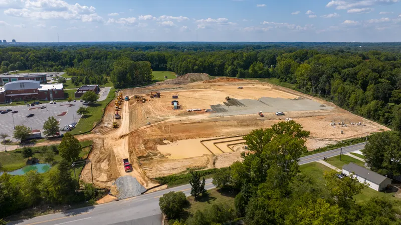 Aerial view of a graded site beside an occupied campus and parking field